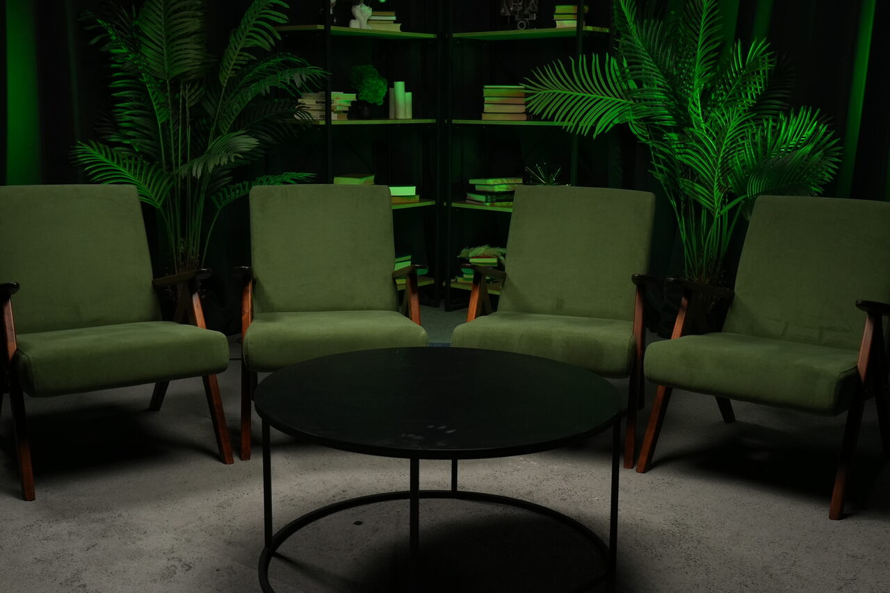 Арендовать Лофт Восторг. Четыре зеленых кресла вокруг круглого черного стола на сером полу. На заднем плане большой стеллаж с книгами и декоративными элементами, зелёные растения. Освещение мягкое, с зелёным оттенком.