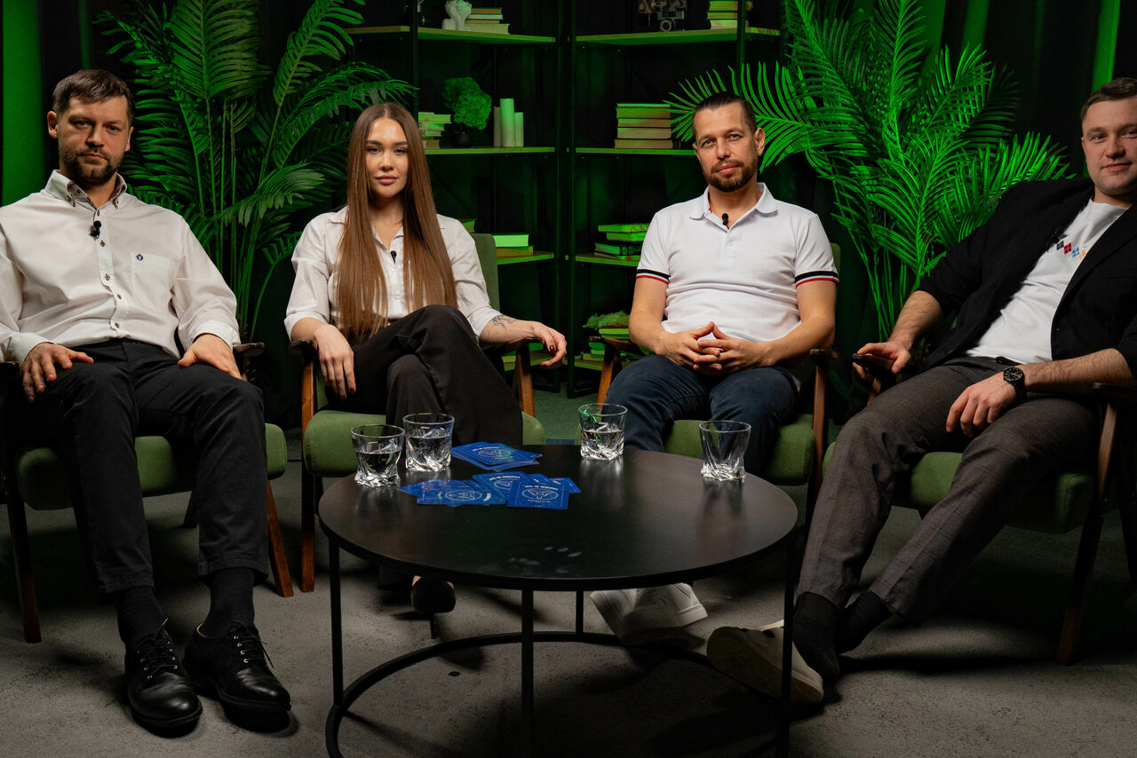 Арендовать Лофт Восторг. Четыре человека сидят на зелёных креслах вокруг круглого чёрного стола, на котором стоят стаканы с водой и лежат синие карточки. Позади видны тёмные стеллажи с книгами и декором, зелёные растения. Стены тёмные. Освещение мягкое.