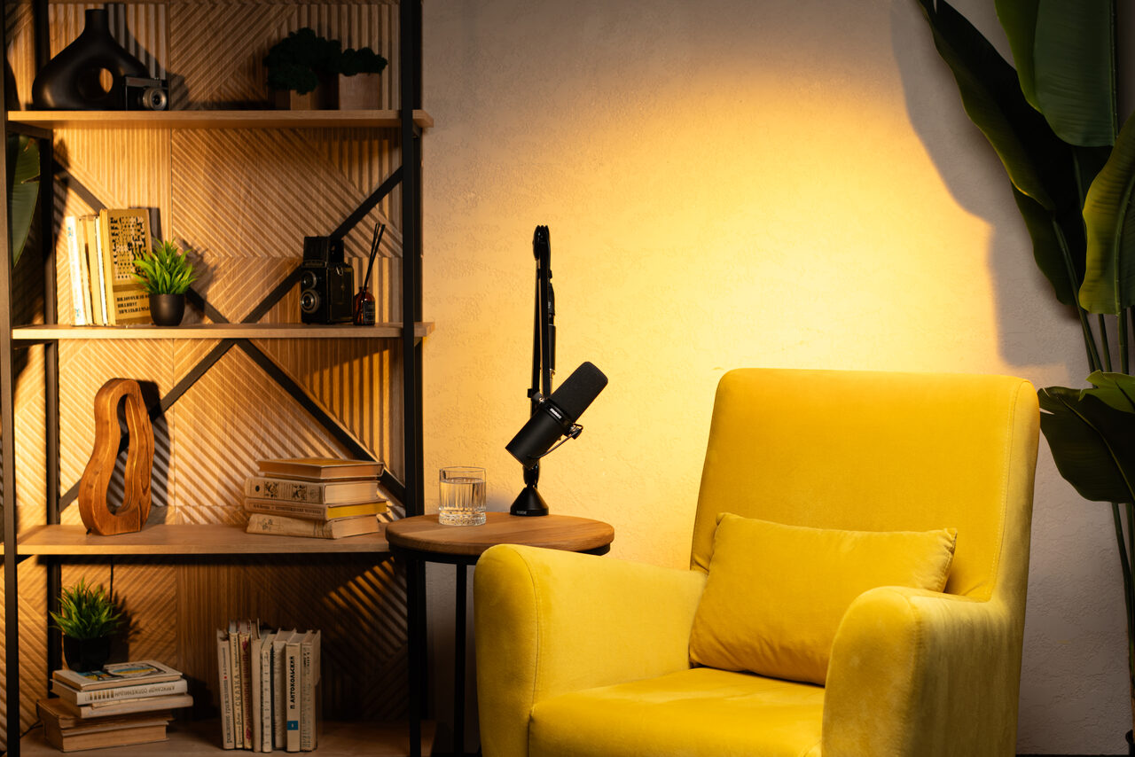 Арендовать Лофт  Эйфория. Комната с жёлтым креслом и подушкой, круглым деревянным столиком с микрофоном и стаканом, стеллажом с книгами и декором, большим зелёным растением. Настенное освещение создаёт тёплый свет, стены и пол спокойных оттенков.