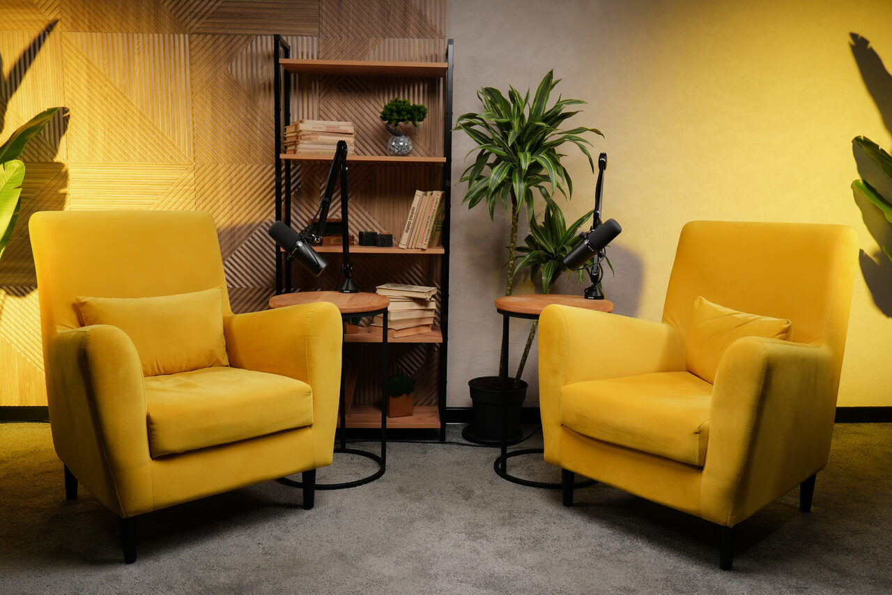 Арендовать Лофт  Эйфория. Желтая комната с двумя креслами, кофейными столиками, микрофонами и книжной полкой. На полке книги и растение. Стены с деревянными панелями и серыми элементами. Растения на полу. Освещение теплое, пол покрыт ковром серого цвета.
