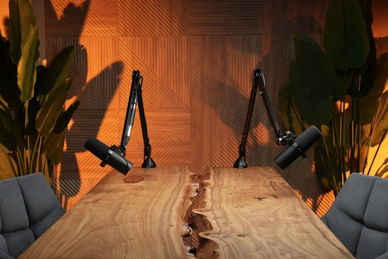 Арендовать Лофт  Эйфория. Деревянный стол с живым краем, два микрофона на стойках, серые кресла. Стена украшена деревянными панелями геометрического узора, зеленые растения по бокам. Теплое освещение, создающее атмосферу уюта и уединения.