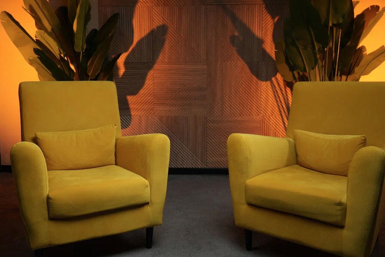 Арендовать Лофт  Эйфория. Две жёлтые кресла с подушками на сером ковровом покрытии. На заднем плане деревянная панель с геометрическим рисунком. По бокам растения с большими листьями. Тёплое освещение создаёт теневые узоры от листвы на стене.