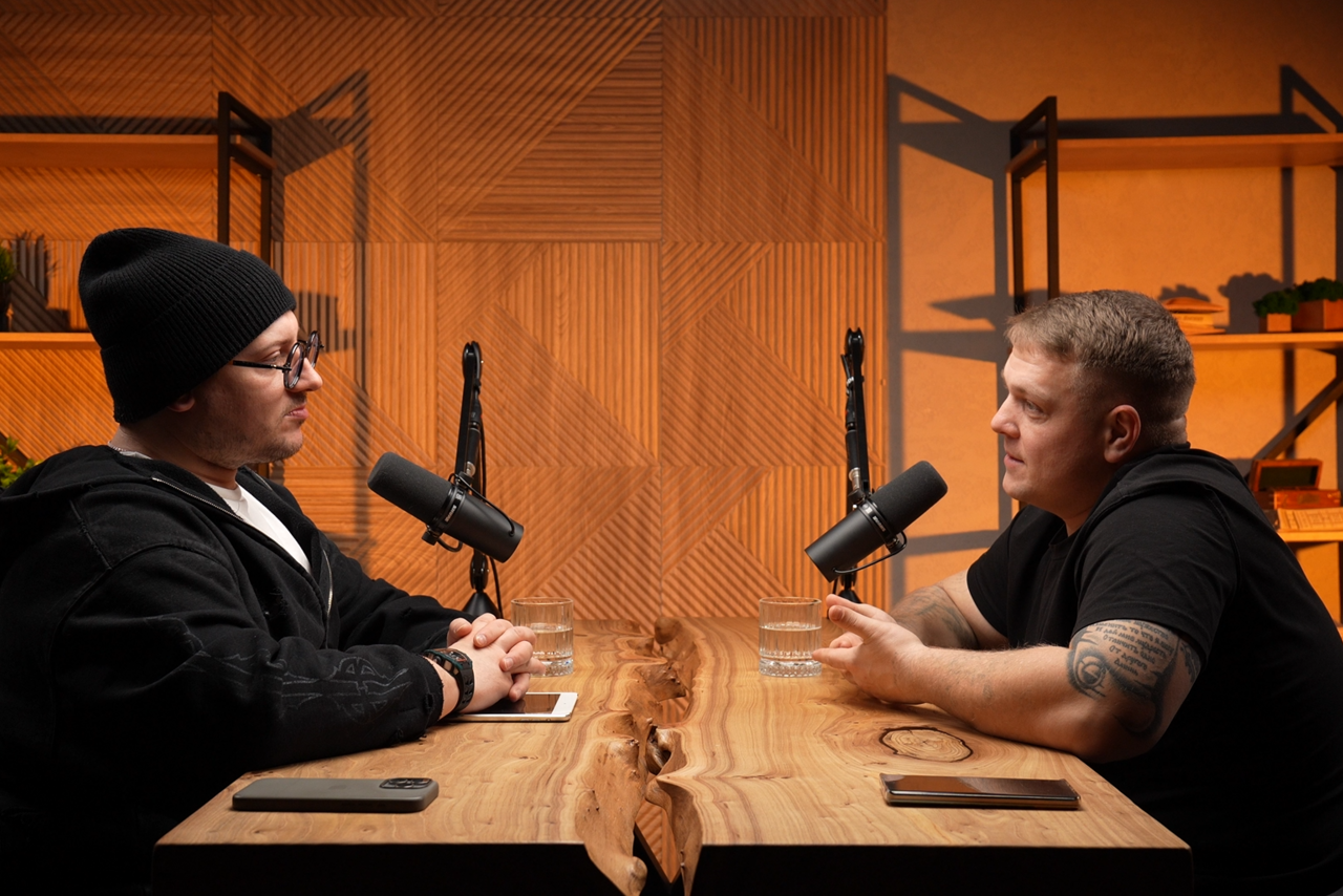 Арендовать Лофт  Эйфория. Студия для записи подкастов, деревянный стол с естественным краем, два микрофона, два смартфона. На столе стаканы с водой. Задняя стенка с геометрическим рисунком, металлические полки сдекором, теплое освещение.