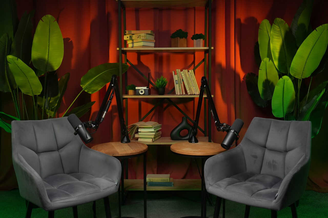 Арендовать Лофт  Эйфория. Две серые кресла и два круглых деревянных стола с микрофонами на стойках. Позади стеллаж с книгами и декором. Красный занавес и зелёные растения по краям. Освещение тёплое, материалы дерева и текстиль, состояние нового.