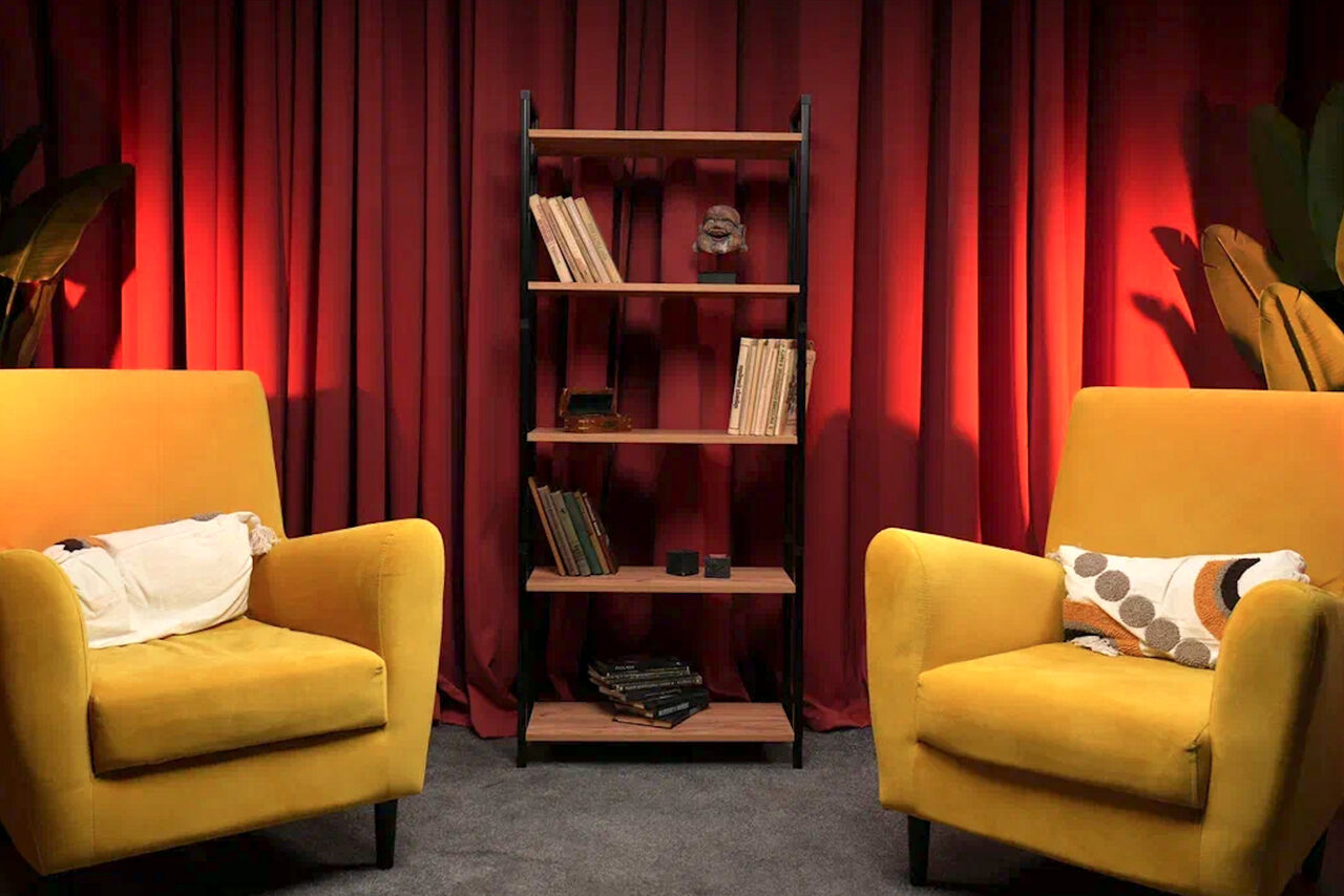 Арендовать Лофт  Эйфория. Жёлтые кресла с декоративными подушками, стеллаж с книгами и предметами на фоне красных штор. Напольное покрытие серое. Освещение мягкое, с акцентом на красные шторы. Слева и справа от кресел крупные зелёные растения.