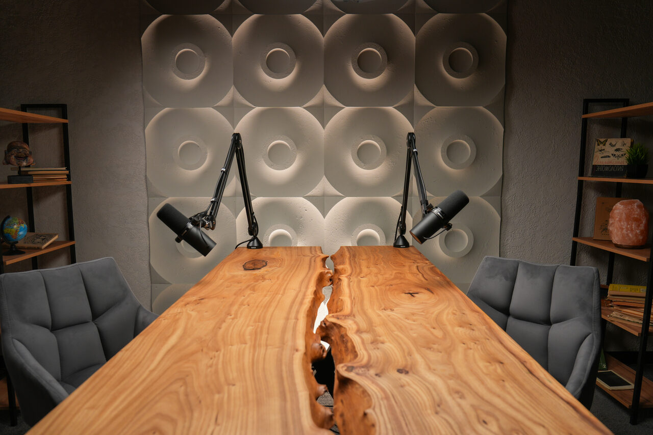 Арендовать Лофт  Эйфория. Деревянный стол с естественной трещиной по центру, четыре серых кресла, два черных микрофона на стойках. Стена с рельефными кругами, две деревянные полки с книгами и декоративными предметами. Теплое освещение, стены серого цвета.