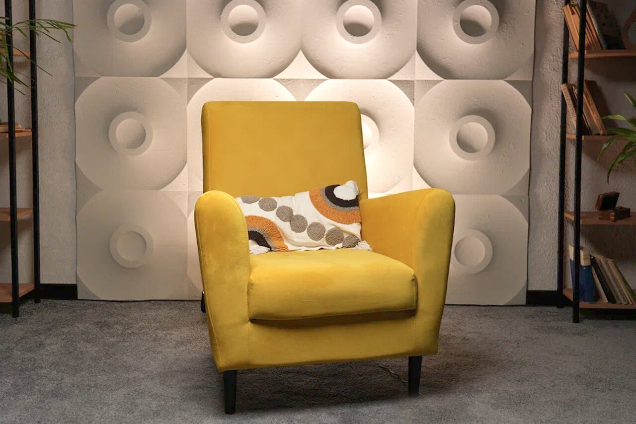 Арендовать Лофт  Эйфория. Жёлтое кресло с подушкой на сером ковре, перед стеной с белыми 3D-панелями круглой формы. По бокам стеллажи с книгами и растениями. Освещение мягкое, равномерное.