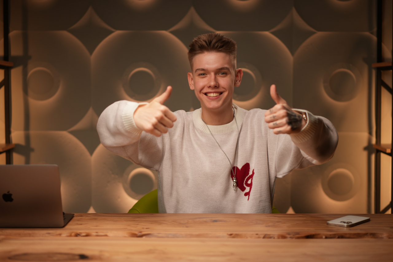 Арендовать Лофт  Эйфория. Офисное пространство с деревянным столом, на столе ноутбук и блокнот. На заднем плане стена с объемным геометрическим узором, освещение мягкое, тёплое. Мужчина в белом свитере показывает жест «большой палец вверх».