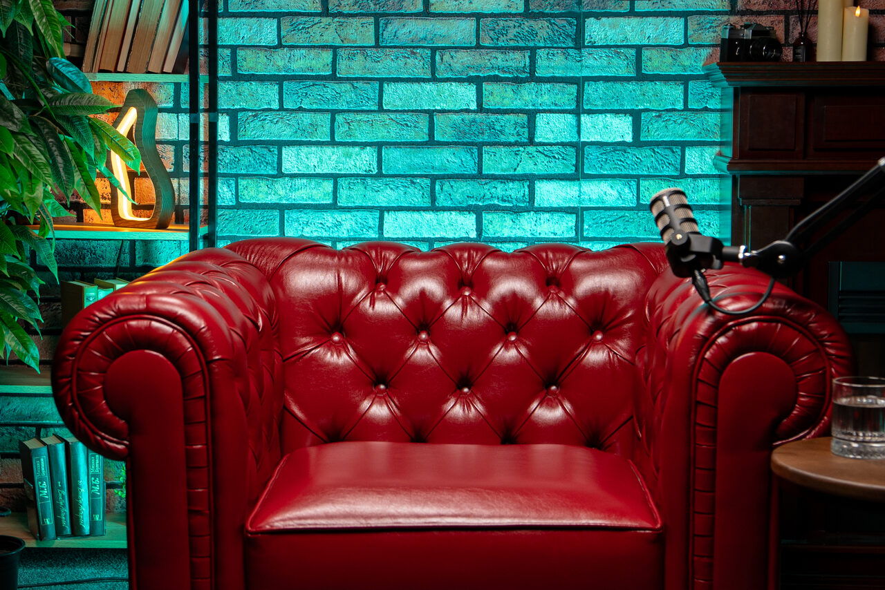 Арендовать Лофт Позитив. Красное кожаное кресло на фоне стены с бирюзовым кирпичным узором. Слева полка с книгами и растением. Справа темно-коричневая стойка с микрофоном на столике и стеклянный стакан. Освещение разноцветное, теплое и яркое.