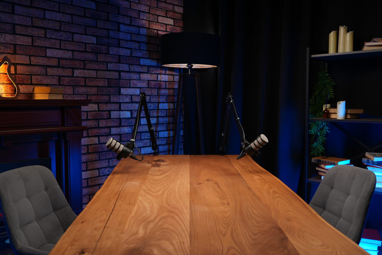 Арендовать Лофт Позитив. Студийное помещение с большим деревянным столом, двумя серыми креслами, микрофонами на стойках. Стена из темного кирпича, черная штора. Полка с книгами, настольная лампа, камин, искусственное освещение с синим оттенком.