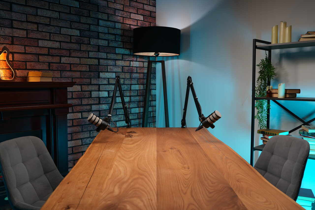 Арендовать Лофт Позитив. Длинный деревянный стол с тремя серыми стульями. На столе два микрофона на стойках. Слева кирпичная стена с камином и лампочкой. Справа стеллаж с книгами и растением. Напольная лампа с чёрным абажуром. Освещение тёплое и неоново-голубое.