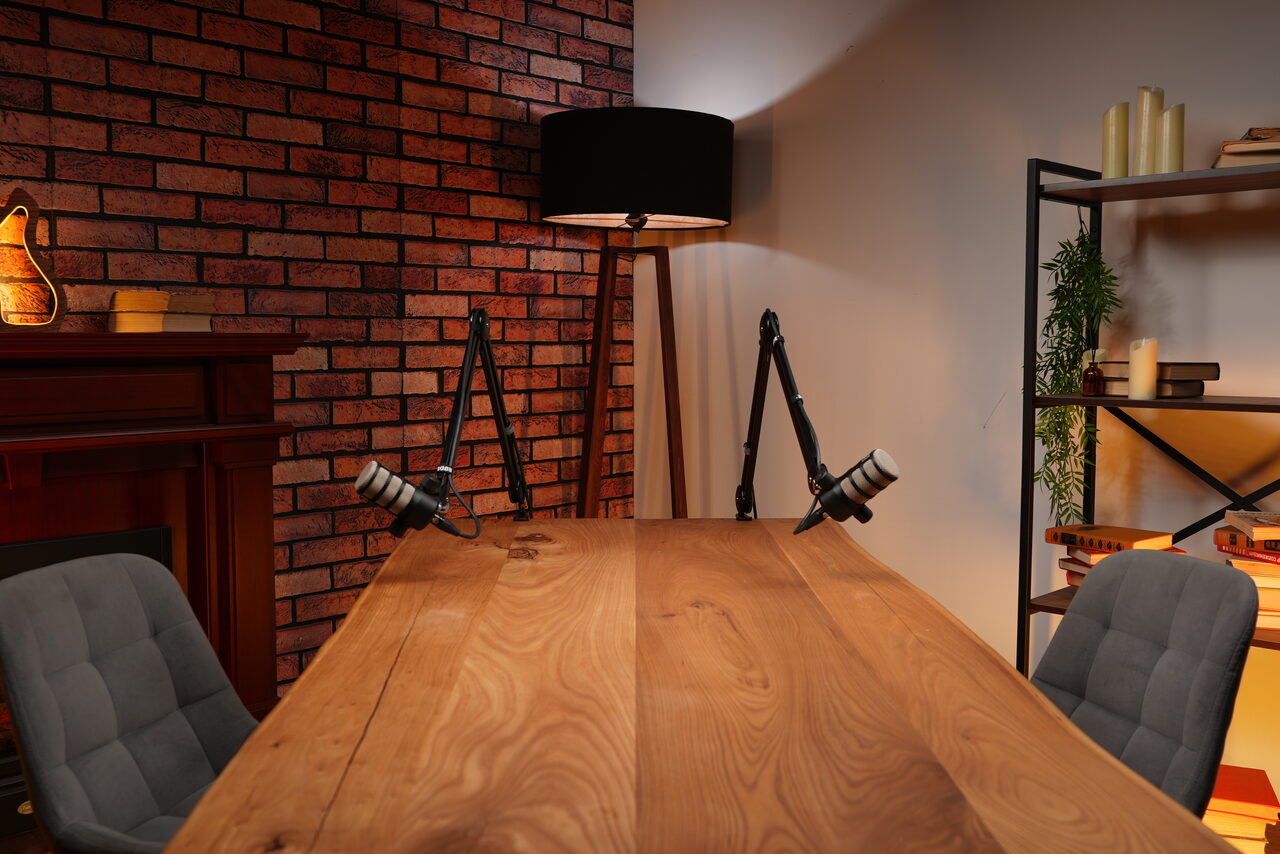 Арендовать Лофт Позитив. Кирпичная стена, длинный деревянный стол, микрофоны на стойках, две серые стулья, напольная лампа с чёрным абажуром, книжная полка с книгами и зеленью, электрокамин, декоративная ваза. Освещение тёплое, стены разного цвета.