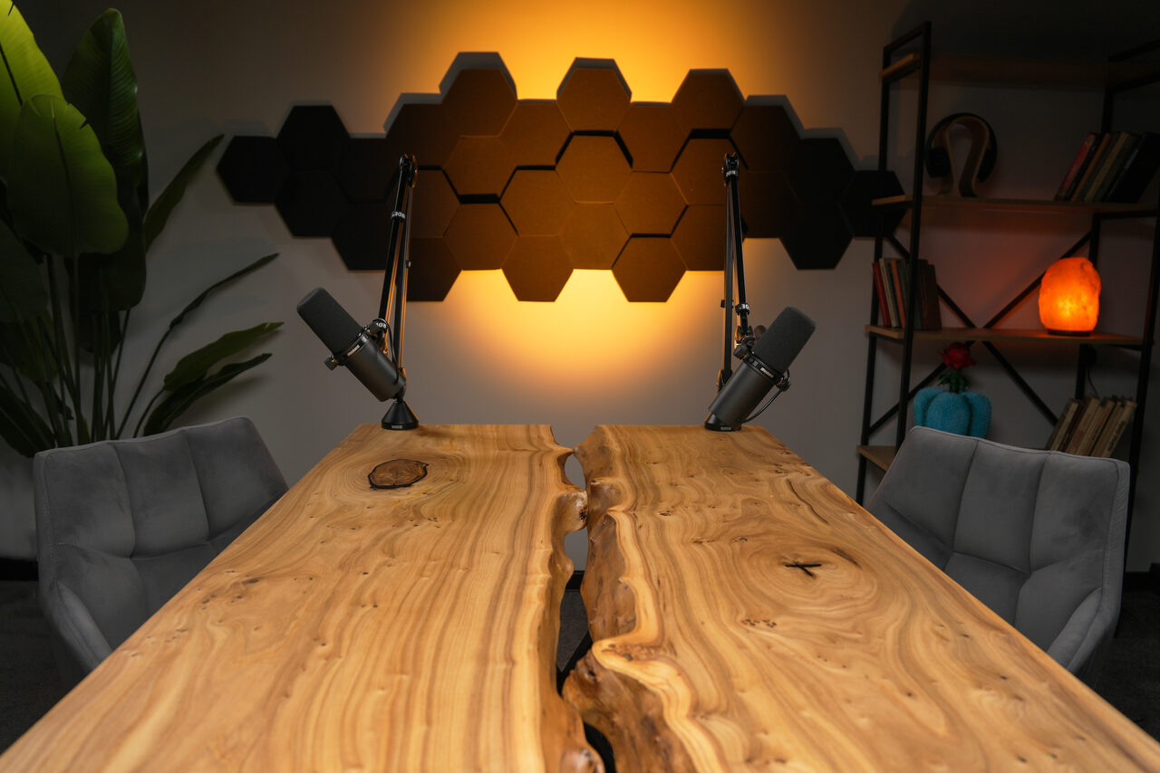 Арендовать Лофт Позитив. Две серые мягкие стулья у стола из светлого дерева с естественным краем и трещиной посередине. Два черных микрофона на длинных стойках. На стене черные шестиугольники и мягкое желтое освещение. Справа черная полка с книгами и декоративными элементами.
