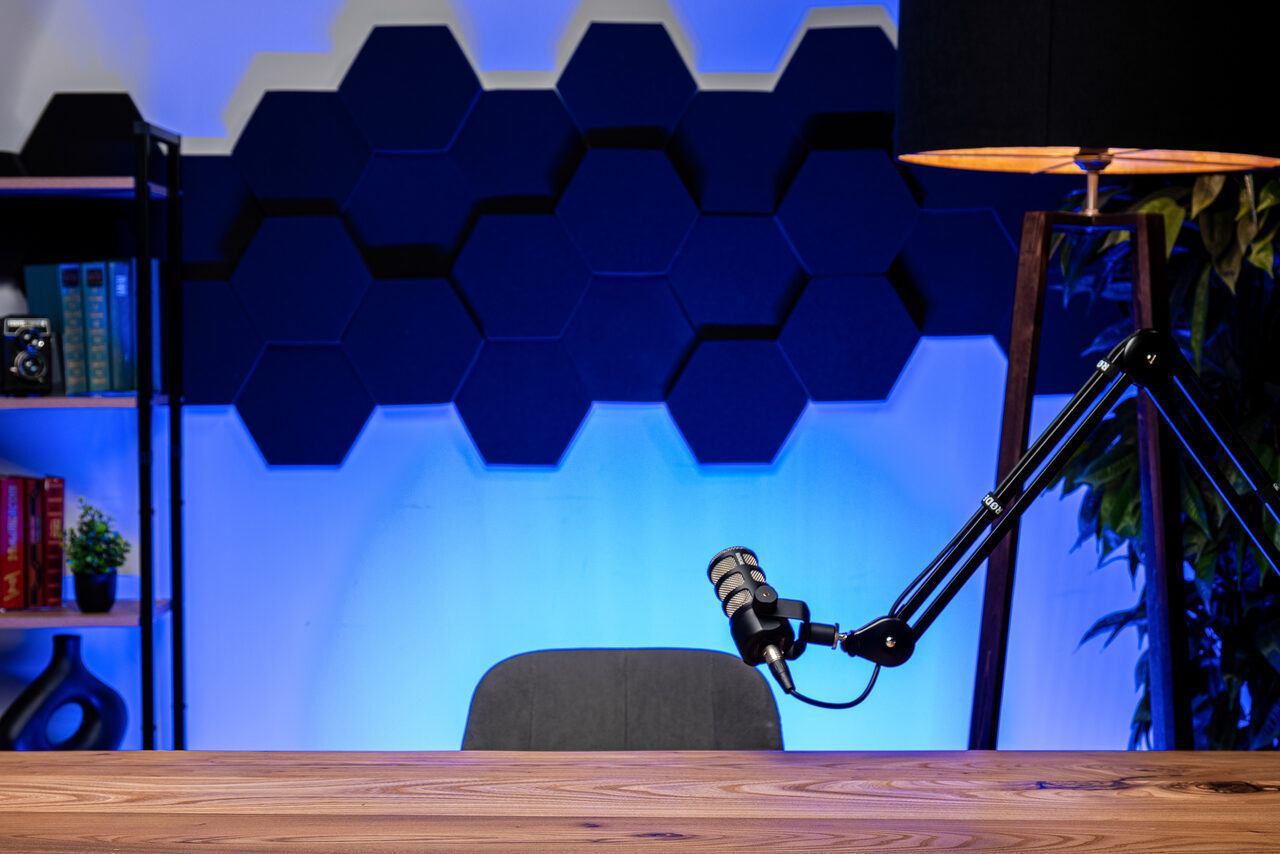 Арендовать Лофт Позитив. Современное помещение с деревянным столом, микрофоном на стойке, чёрным стулом. На стене синие шестиугольные панели, в углу книжные полки с декоративными элементами. Напольная лампа с абажуром, мягкое освещение.