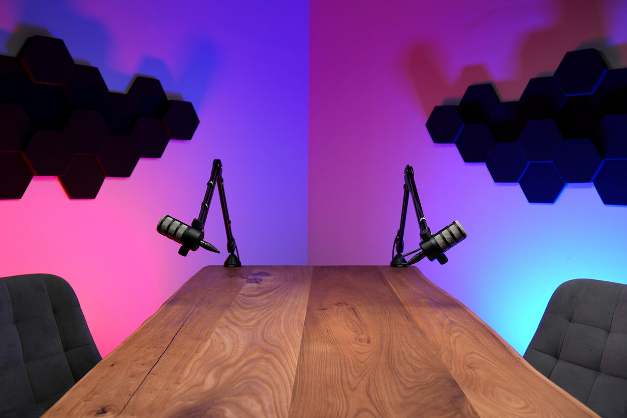 Арендовать Лофт Позитив. Деревянный стол с двумя микрофонами на стойках, по обе стороны находятся стулья тёмного цвета. На стене декоративные элементы в виде чёрных шестиугольников. Освещение неоновое, розовый и синий цвета.