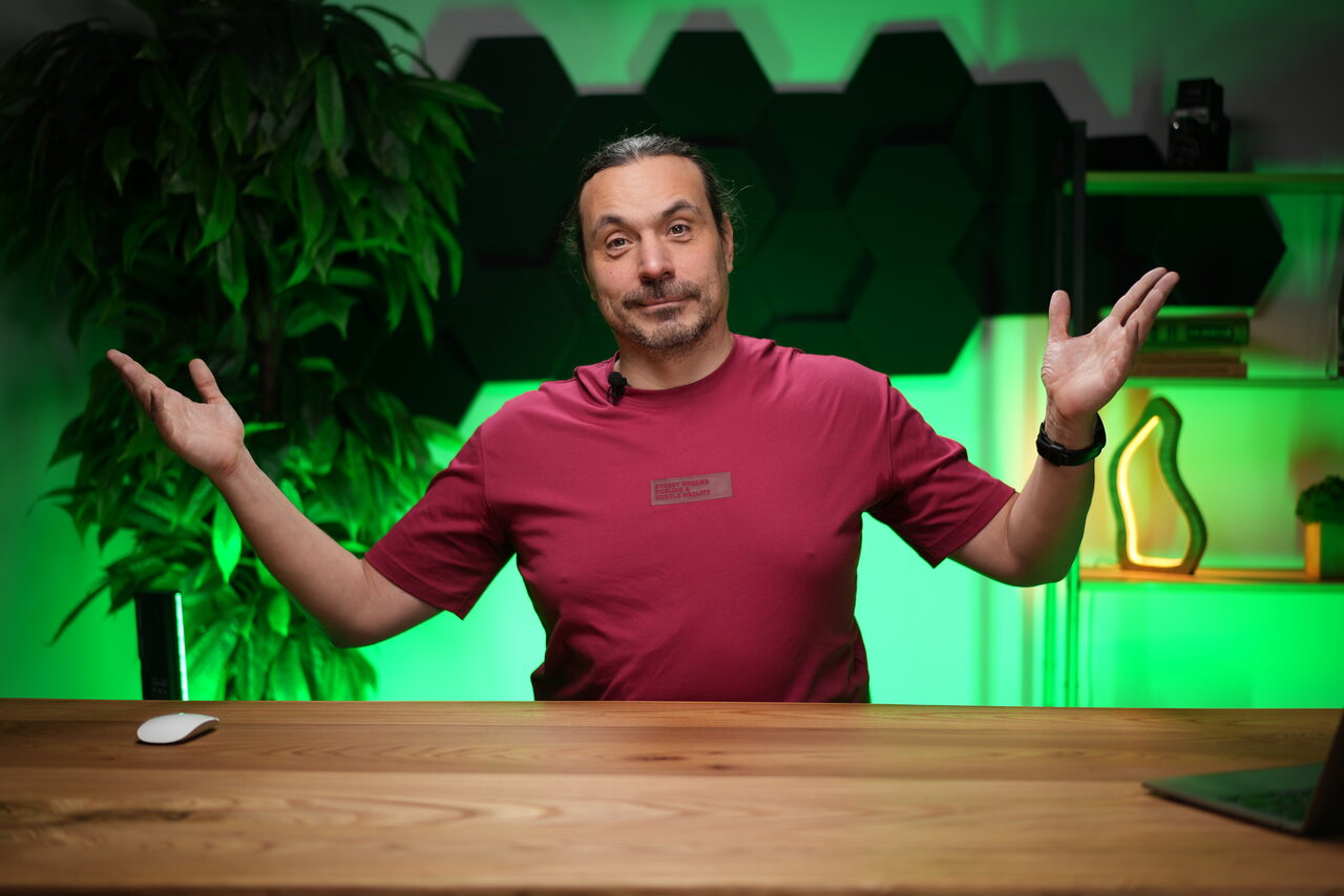 Арендовать Лофт Позитив. Человек в красной футболке сидит за деревянным столом. На фоне темно-зеленые панели, зелёная подсветка, полки с книгами и растением, искусственное растение слева. Справа на столе мышь, частично виден ноутбук.