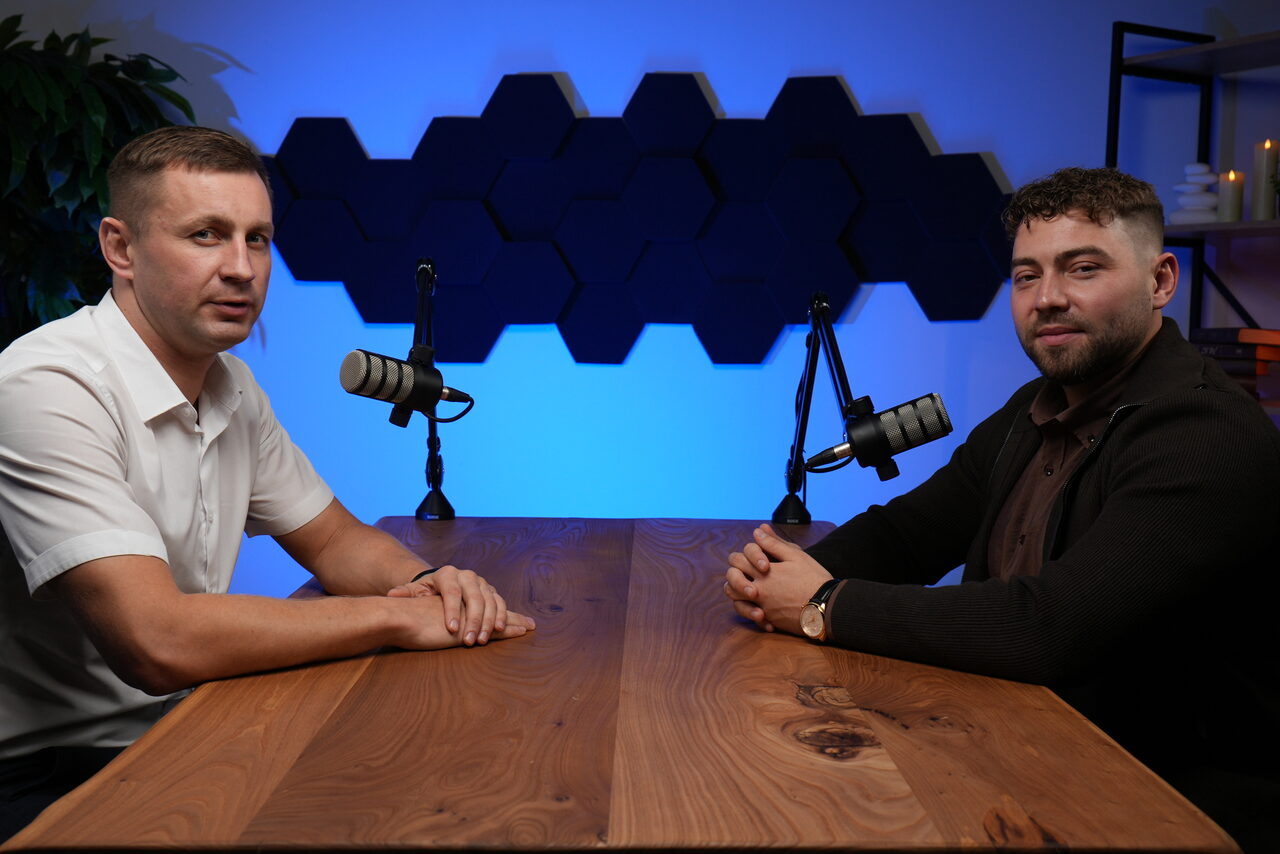 Арендовать Лофт Позитив. Помещение с деревянным столом, двумя микрофонами на стойках, сидят два человека. Синее акцентное освещение. На стене чёрные шестиугольные панели, справа полка с декором и свечой. Слева растение.