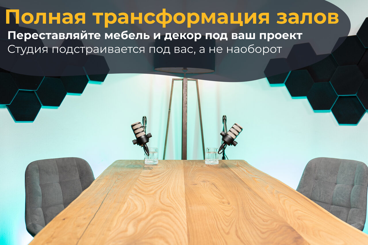 Арендовать Лофт Позитив. Пространство со столом из светлого дерева и двумя серыми стульями. На столе находятся два микрофона на стойках. Светлая стена с черными шестиугольными панелями. Потолочная лампа с тремя лампочками. Текст на изображении сверху.