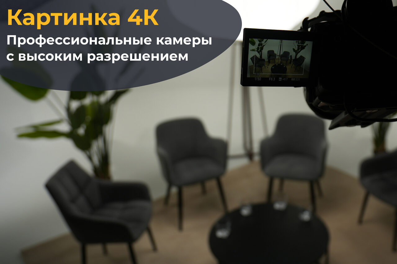 Арендовать Лофт Циклорама. Студийное пространство с четырьмя серыми креслами вокруг круглого черного стола, на котором стоят три стакана воды. Съемочная камера на штативе располагается в правой части кадра. Светлые стены и растения на фоне. Текст на изображении: «Картинка 4К. Профессиональные камеры с высоким разрешением».