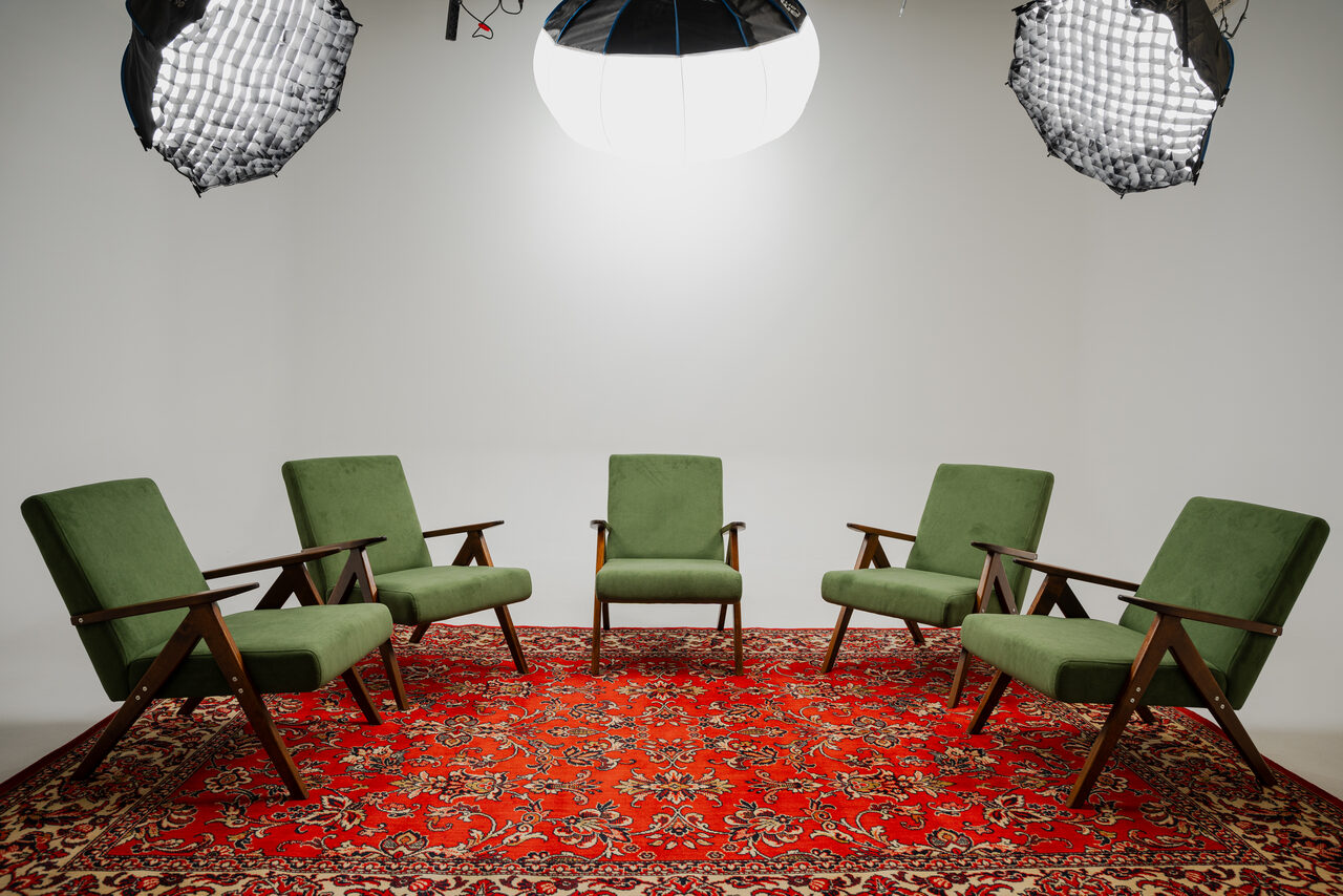 Арендовать Лофт Циклорама. Круговая расстановка зеленых кресел с деревянными подлокотниками на красном ковре с узором; белые стены; два больших осветительных прибора вверху.