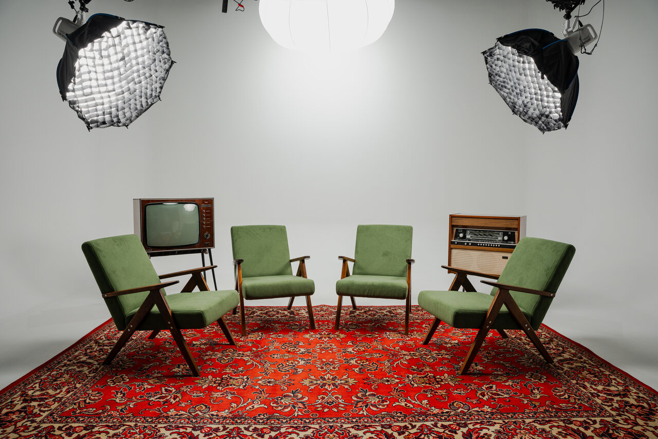 Арендовать Лофт Циклорама. Студийное пространство с двумя студийными софтбоксами, тремя зелеными креслами, ретро-телевизором и радиоприемником на фоне белой стены. Пол украшает красный восточный ковер с узором.