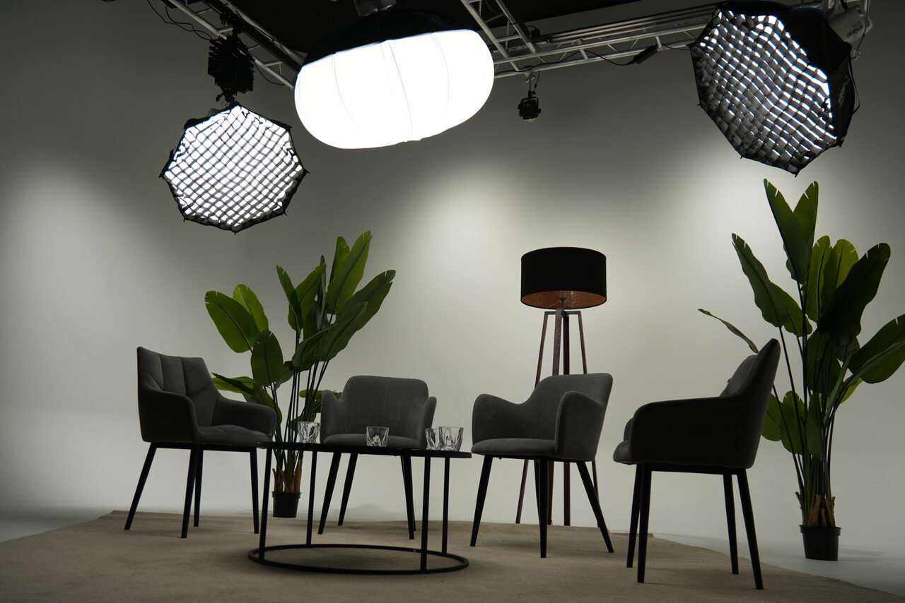Арендовать Лофт Циклорама. Студийное пространство с серыми стенами, четыре тёмно-серых кресла вокруг чёрного круглого стола, стеклянные стаканы, напольная лампа на трёх ножках, несколько крупных зелёных растений, светло-серый ковёр, три крупных осветительных прибора сверху.