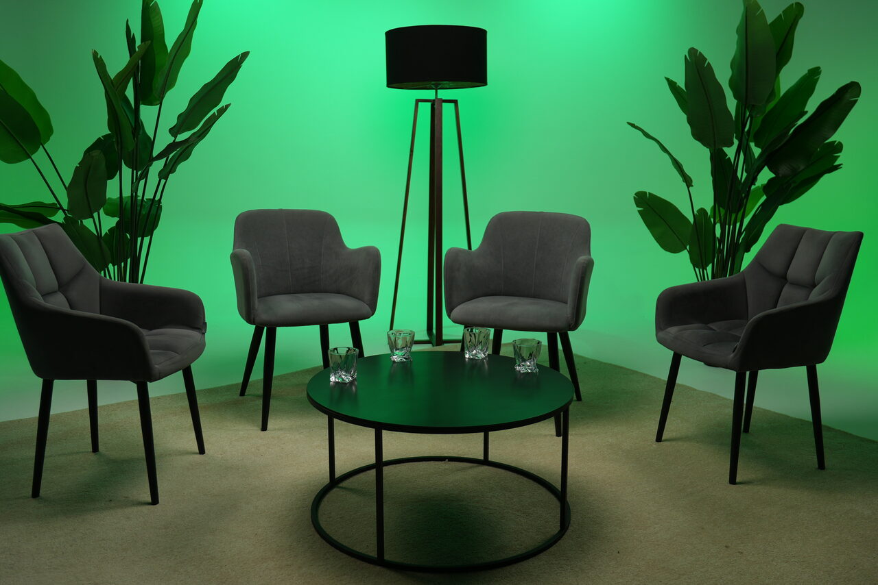 Арендовать Лофт Циклорама. Современное пространство с четырьмя серыми креслами вокруг круглого черного стола. На столе четыре стакана. Стоячая лампа с черным абажуром. Зеленая подсветка стен. Растения по углам. Пол ковровый, бежевого цвета.