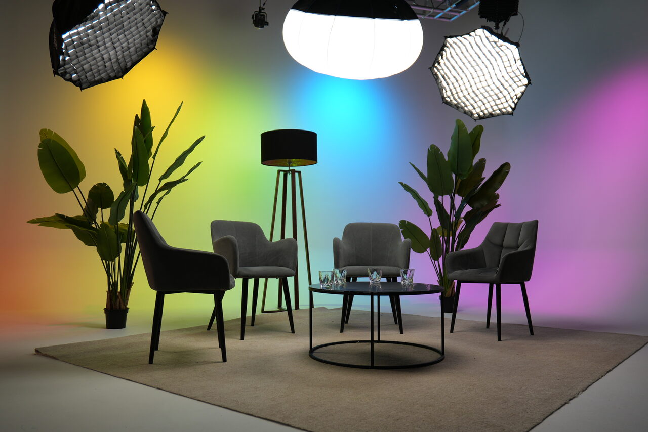 Арендовать Лофт Циклорама. Съемочная студия с четырьмя серыми креслами и круглым стеклянным столом на светлом ковре. Растения в горшках расположены вокруг зоны. Оборудование для освещения с разноцветной подсветкой на стене. Лампы на штативах и висящие светильники.