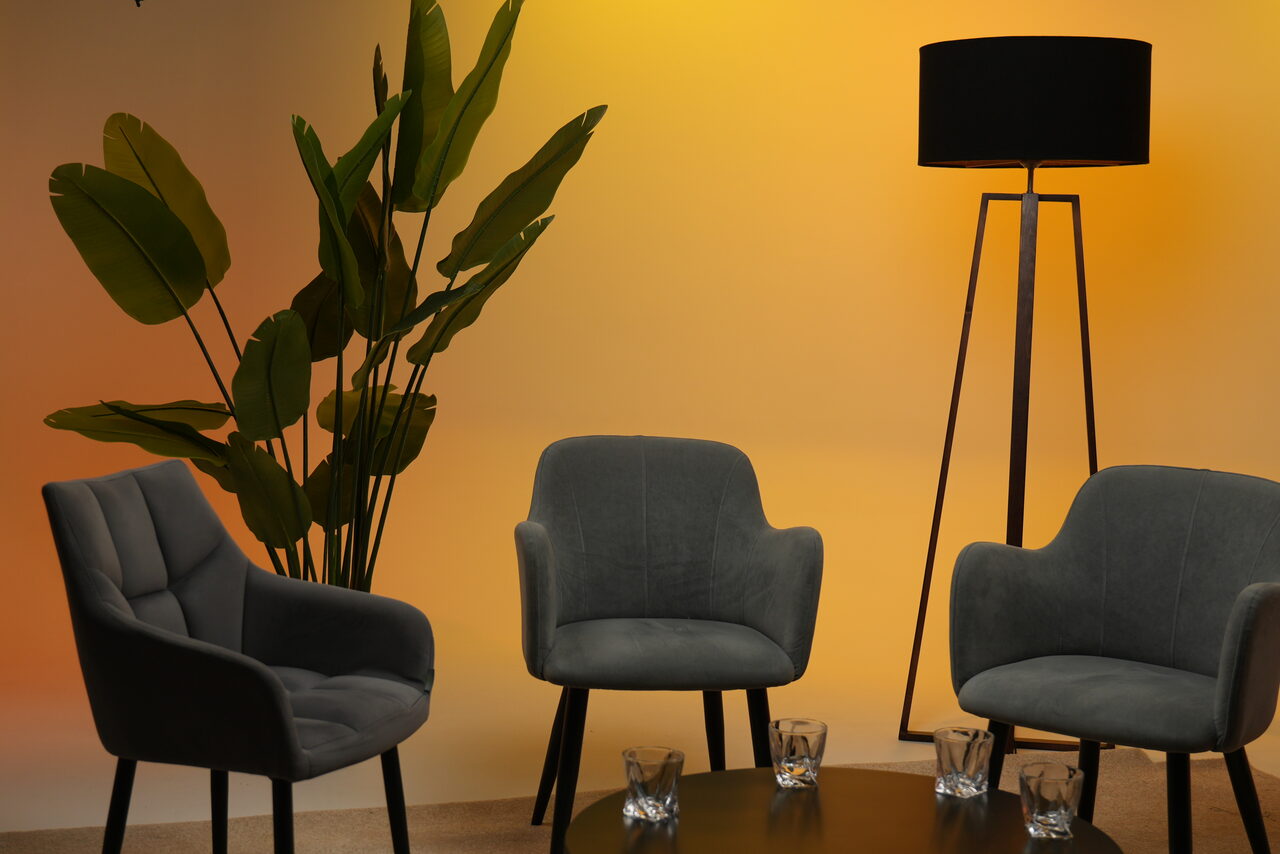 Арендовать Лофт Циклорама. Современное помещение с тремя серыми креслами, черным столом с четырьмя стеклянными стаканами, высоким черным торшером и искусственными зелеными растениями. Фон с градиентом от желтого к светло-коричневому.