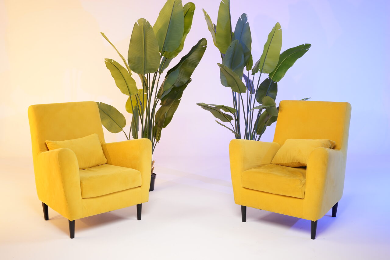 Арендовать Лофт Циклорама. Два желтых кресла с подушками, расположенных напротив друг друга, рядом с зелеными растениями. Пол белый, освещение разноцветное, с оттенками желтого и синего. Кресла имеют черные ножки.