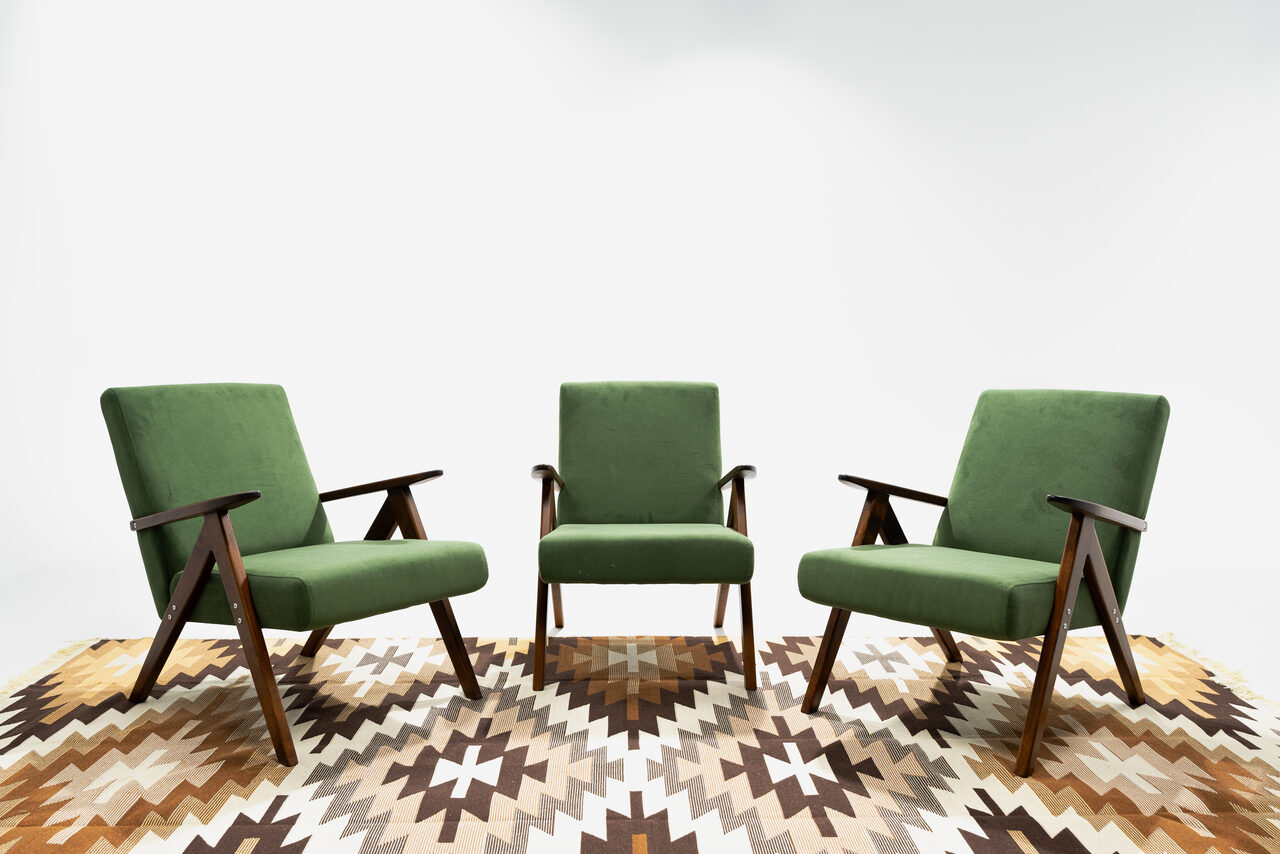 Арендовать Лофт Циклорама. Три зеленых кресла с деревянными подлокотниками расположены полукругом на ковре с геометрическим узором в коричневых и бежевых тонах. Фон белый.