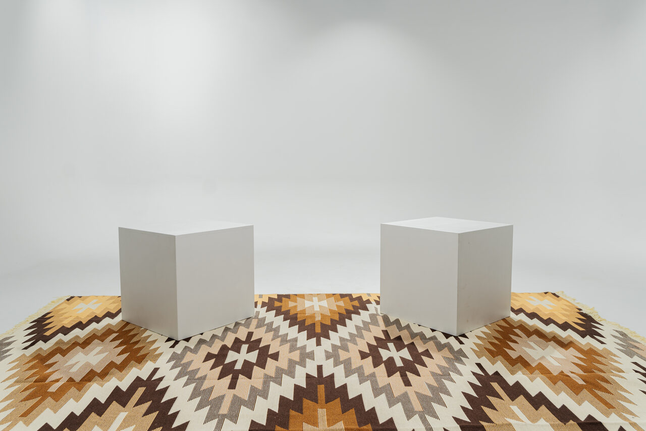 Арендовать Лофт Циклорама. Белая комната с двумя белыми кубами на полу. Пол покрыт ковром с геометрическим узором в коричневых и бежевых тонах. Стены и потолок белые, несколько источников встроенного освещения.