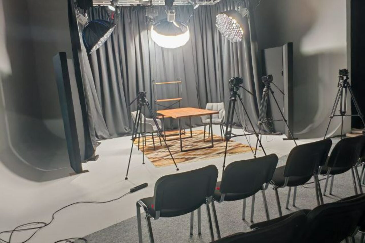 Арендовать Лофт Циклорама. Студийное пространство с черными шторами, круглыми светильниками и тремя камерами на штативах. Деревянный стол с двумя стульями на ковре. В переднем плане ряд черных стульев. Белый пол, серая стена.