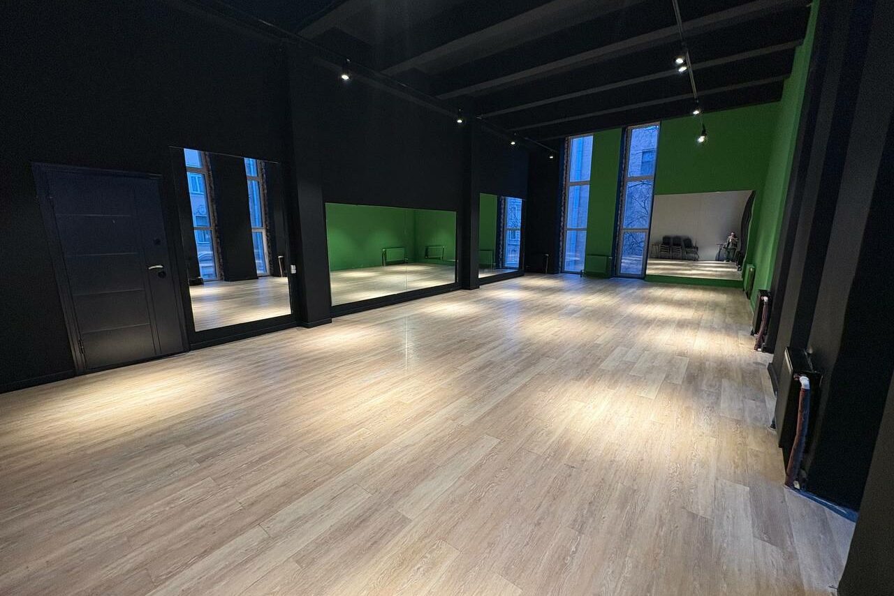Арендовать Танцевальный зал 80 м.кв.. Просторное помещение с деревянным полом, черными стенами и зелеными акцентами. Зеркальная стена слева, высокие окна справа и на задней стене. Черная дверь слева. На заднем плане стулья, освещение с потолка.