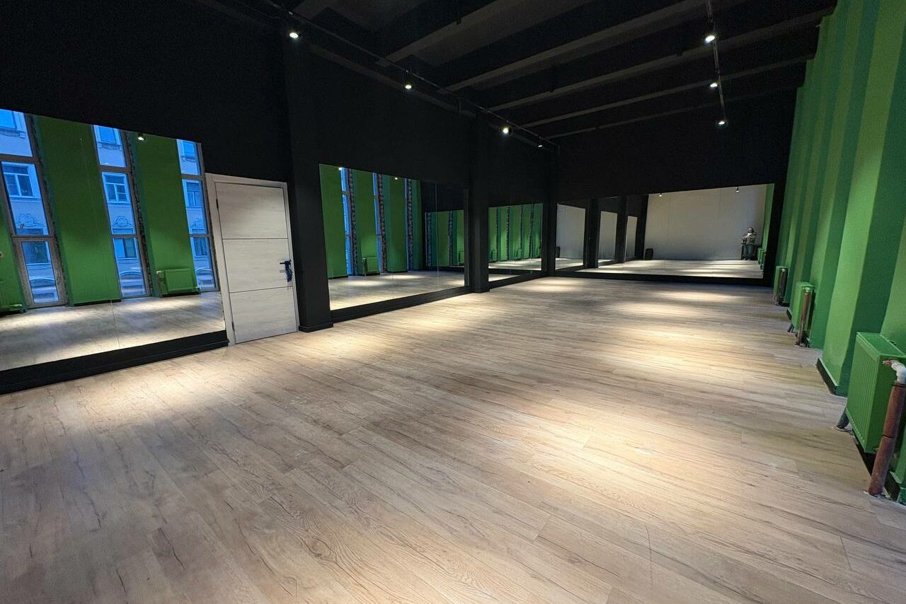 Арендовать Танцевальный зал 80 м.кв.. Просторное помещение с деревянным полом, стены в зелёных и тёмных оттенках, большие окна с отражением домов, светодиодное освещение на потолке, белая дверь сбоку.