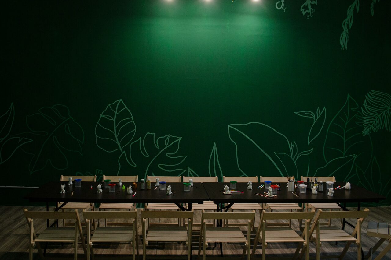 Арендовать Хендмейдщики. Лофт в лесной тематике. Пространство с длинным черным столом и деревянными стульями, расположенными в ряд. На столе множество канцелярских принадлежностей. Задняя стена зеленая с тонкими рисунками листьев. Потолочное освещение с тремя светильниками. Пол светлый.