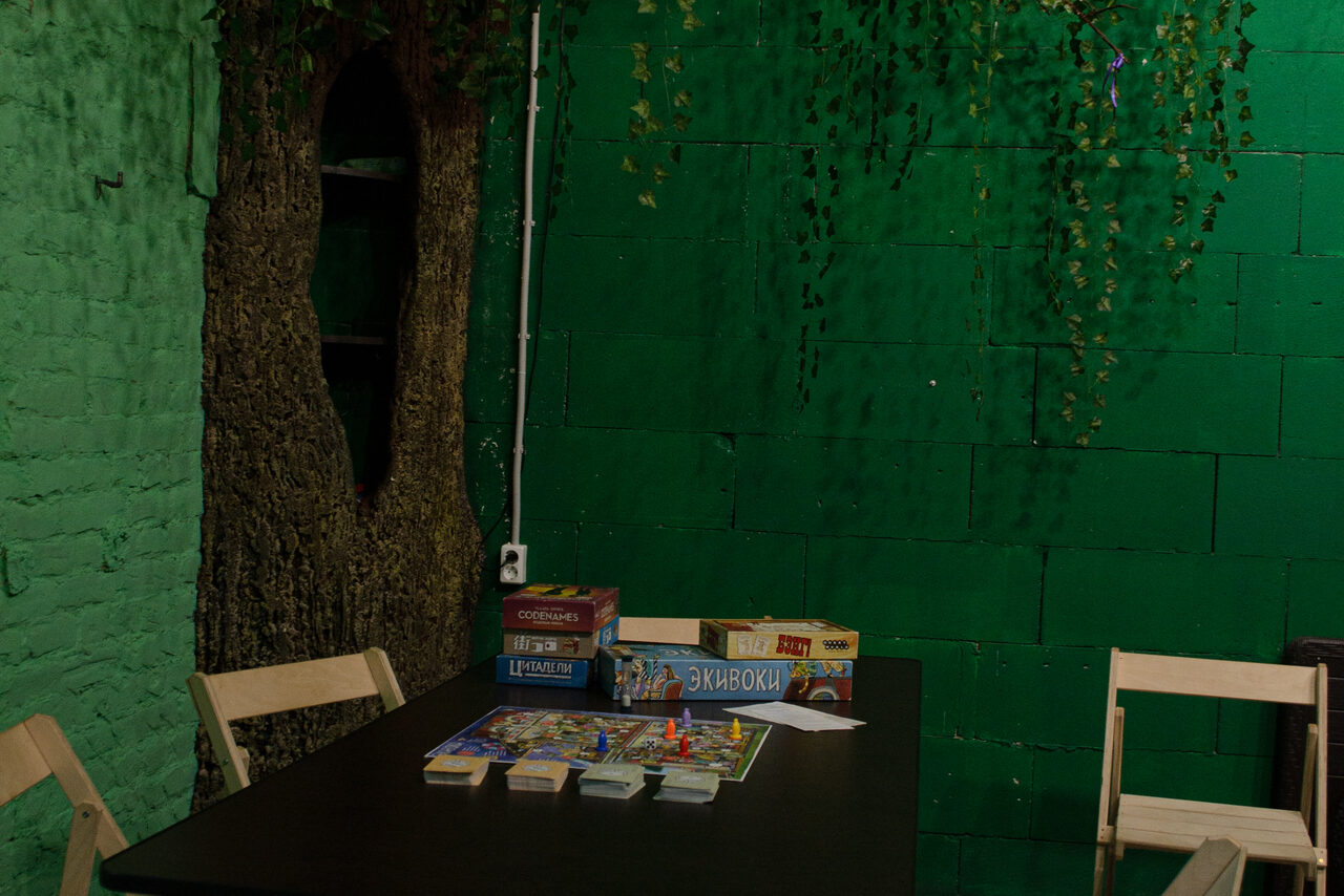Арендовать Хендмейдщики. Лофт в лесной тематике. Игровое пространство с черным столом и деревянными стульями. На столе настольные игры и карты. Зеленая кирпичная стена, декорированная искусственными растениями. Дерево с полками на заднем фоне. Электрическая розетка на стене.