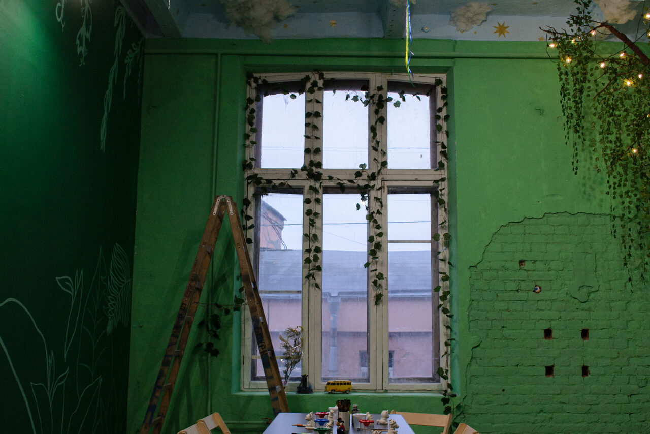 Арендовать Хендмейдщики. Лофт в лесной тематике. Зеленая комната с высоким окном, украшенным искусственными растениями. Стены окрашены в зеленый, частично с обнаженной кирпичной кладкой. В центре деревянный стол с предметами. Деревянная лестница, потолок светло-голубой с облаками и звездами.