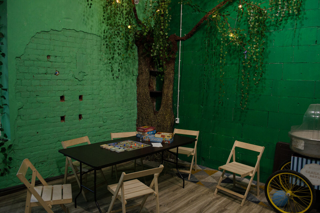 Арендовать Хендмейдщики. Лофт в лесной тематике. Пространство с зелеными кирпичными стенами, декорированное искусственным деревом и гирляндами. Шесть деревянных стульев вокруг прямоугольного черного стола с настольными играми. Пол из ламината светлого оттенка.