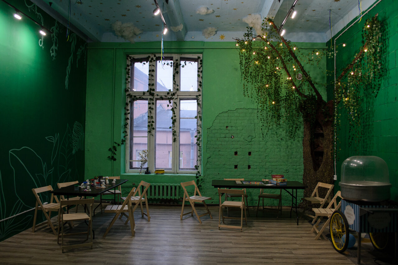 Арендовать Хендмейдщики. Лофт в лесной тематике. Просторное помещение с тёмно-зелёными стенами и деревянным полом. Деревянные складные стулья расставлены вокруг чёрных столов. На стене декоративное дерево с гирляндой. Большое окно с растениями, радиатором. Слева от окна аппарат с прозрачной крышкой. Подвесные светильники.