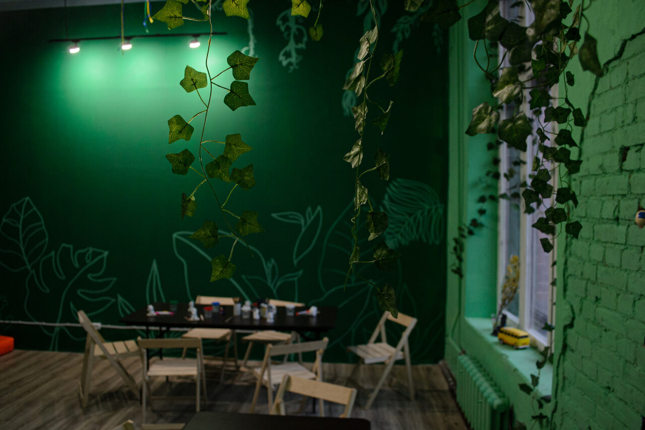 Арендовать Хендмейдщики. Лофт в лесной тематике. Комната с зелеными стенами и рисунками листьев. Деревянные складные стулья, стол с книгами и коробками. Подвесной светильник с тремя лампами. Паркетный пол, кирпичная стена, зелёные растения свисают с потолка. Радиатор под окном.
