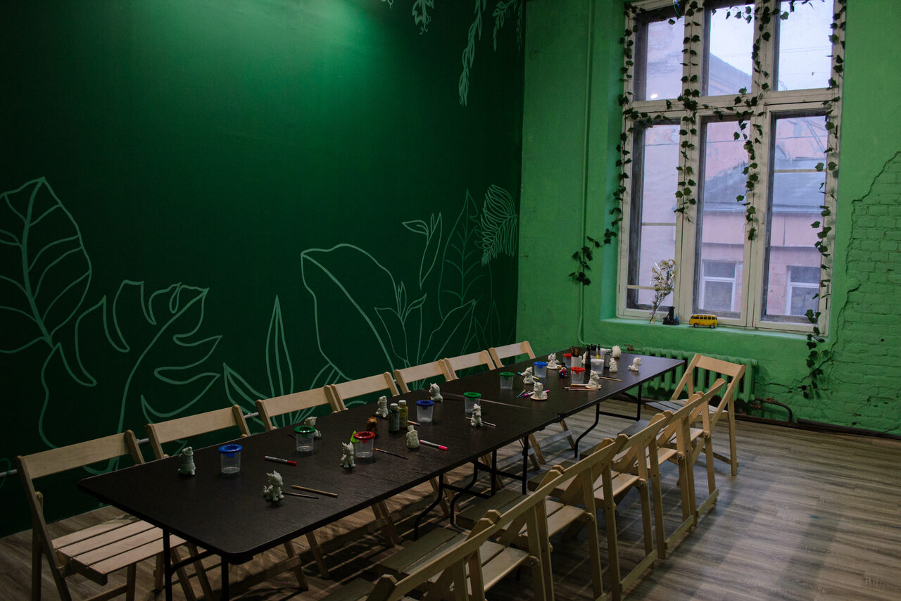 Арендовать Хендмейдщики. Лофт в лесной тематике. Просторное помещение с зеленой стеной, на которой нарисованы контуры листьев. Длинный черный стол окружен деревянными стульями. На столе банки с красками и кисти. На окне свисают растения. Пол светлый, деревянный.