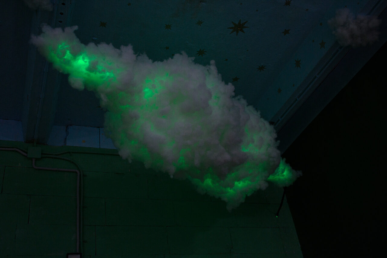 Арендовать Хендмейдщики. Лофт в лесной тематике. Облако из искусственного материала с зелёной подсветкой на фоне стены мятного цвета и потолка с изображением звёзд. Видны трубы или кабели вдоль стены. Тёмное освещение.