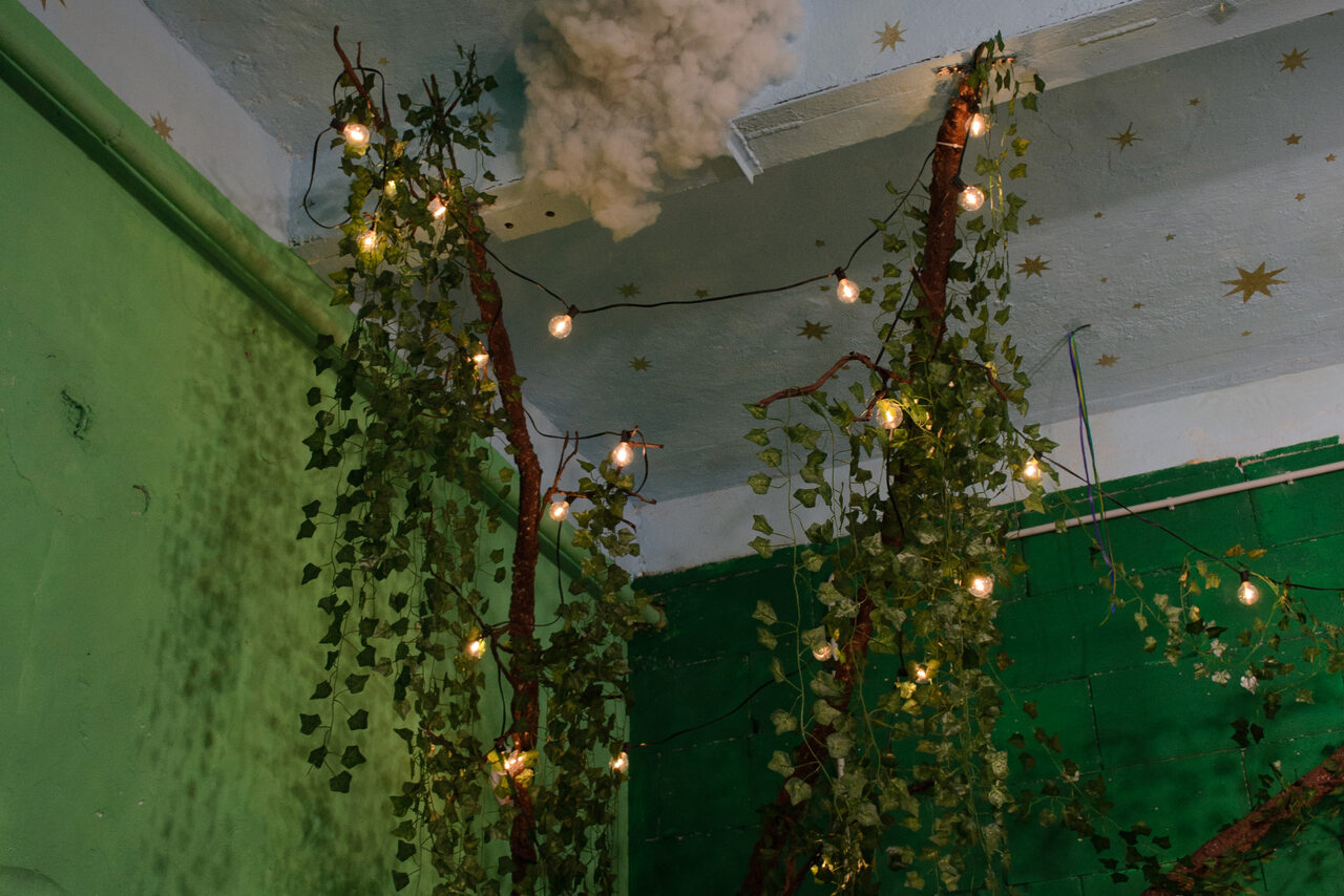 Арендовать Хендмейдщики. Лофт в лесной тематике. Зелёные стены с искусственными лианами и маленькими лампочками на потолке, украшенном звёздами. Потолок белого цвета, на нём видны текстурные элементы, напоминающие облака. Состояние хорошее, освещение атмосферное.
