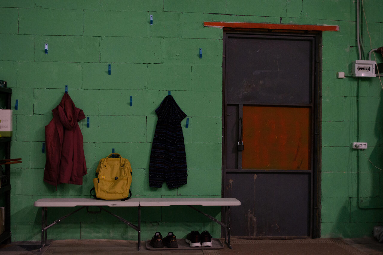 Арендовать Хендмейдщики. Лофт в лесной тематике. Зеленая кирпичная стена с металлической дверью, обрамленной красной полосой. На стене синие крючки с одеждой и рюкзаком. Спереди складной белый стол с обувью. На стене виден светильник и электрическая проводка.