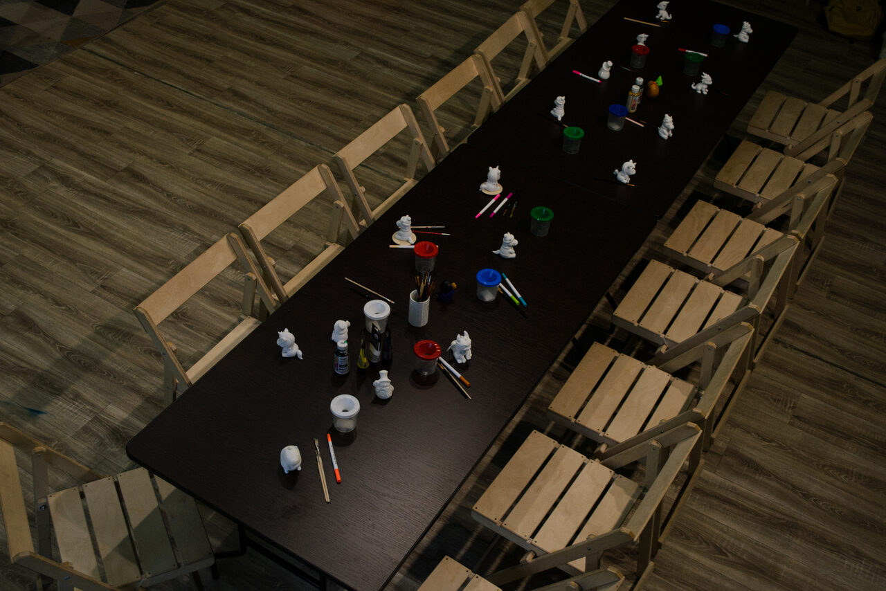 Арендовать Хендмейдщики. Лофт в лесной тематике. Длинный темный стол с деревянными складными стульями вокруг. На столе размещены краски, кисти и небольшие фигурки. Пол деревянный. Освещение приглушенное.