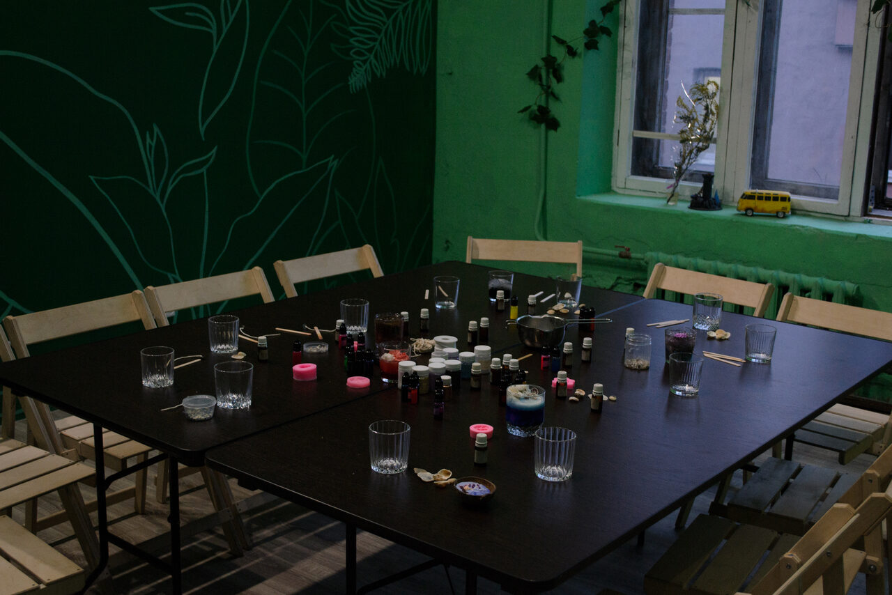 Арендовать Хендмейдщики. Лофт в лесной тематике. Пространство с большим тёмным столом, окружённым деревянными стульями. На столе расположены банки и стаканы. На зелёной стене рисунок с растительным узором. Подоконник с жёлтой игрушкой и растением. Естественное освещение из окна.