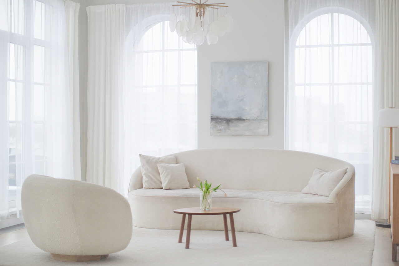 Арендовать Mimika(Мимика). Просторная фотостудия. Светлая комната с большими арочными окнами. Белый диван с подушками, рядом кофейный столик с вазой и цветами. Белое кресло, картина на стене. Пол покрыт светлым ковром. Потолок с белой люстрой. Шторы на окнах.