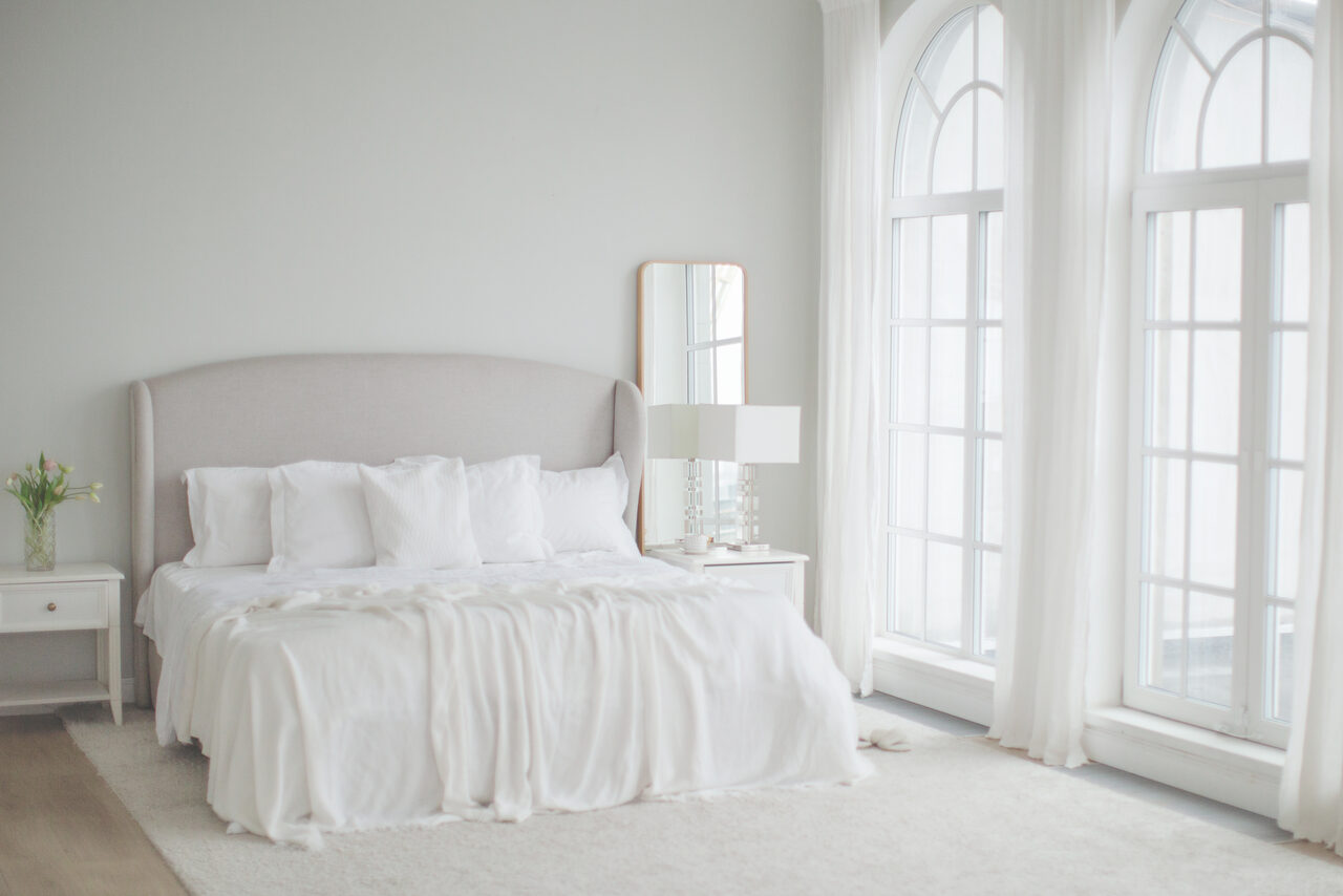 Арендовать Mimika(Мимика). Просторная фотостудия. Спальня с большой кроватью и белым постельным бельём, светло-серые стены, два высоких окна с арочными вставками, зеркало в раме, прикроватный столик с лампой, светлый ковёр, ваза с цветами, мягкое естественное освещение.