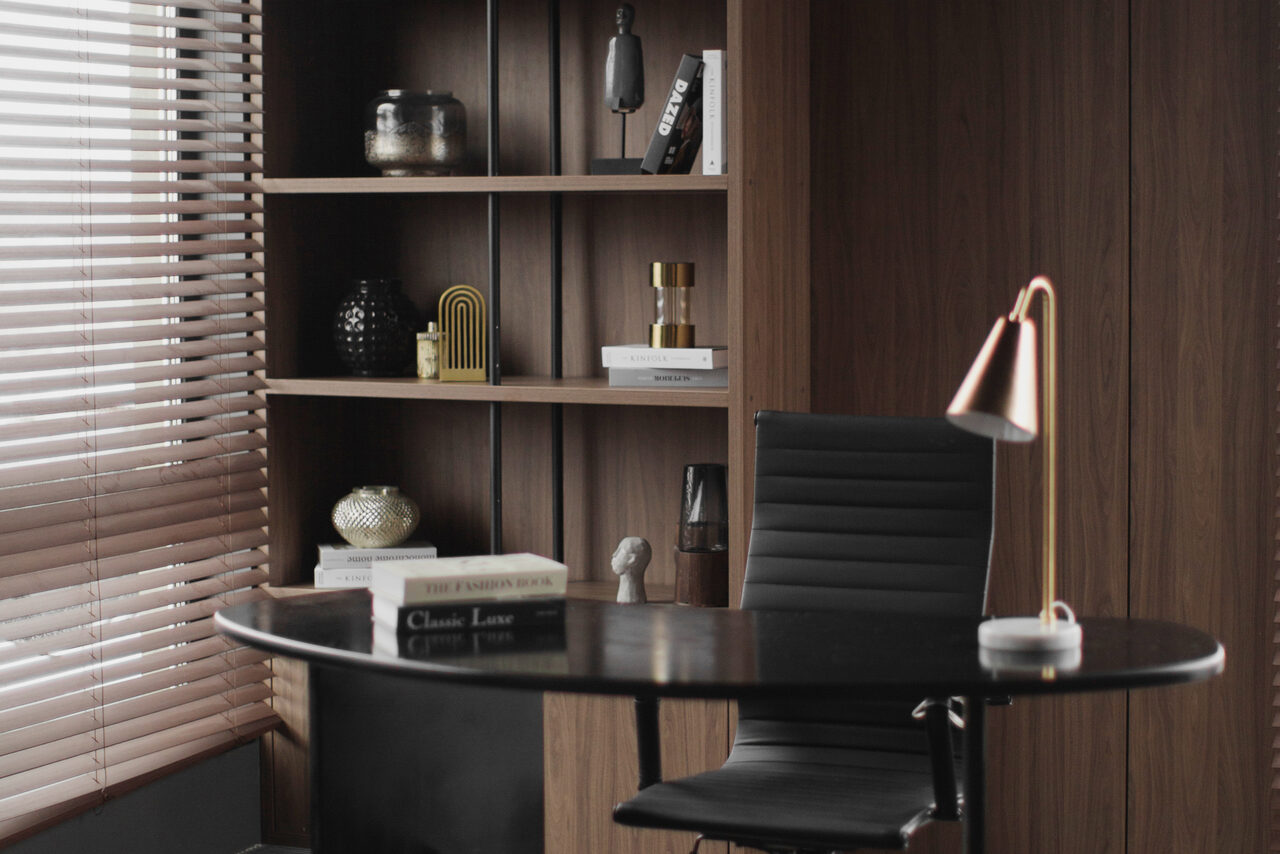Арендовать Mimika(Мимика). Просторная фотостудия. Современный офис с темной деревянной отделкой. Круглый стол с черной столешницей и черным креслом. На столе медная лампа и книга. На заднем плане полки с декором и книгами. Горизонтальные жалюзи на окнах.